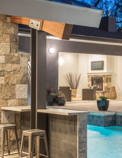 Outdoor patio with a stone bar, stools, and a turquoise lit pool beside a modern home with large windows and trees in the background.