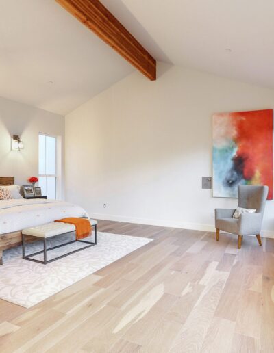 Modern bedroom with a large bed, two side tables, a colorful abstract painting, and an armchair. Light wood floors and a wooden ceiling beam complete the decor.