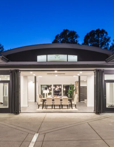 Modern house exterior with large open sliding doors revealing a dining area. The building is dark in color with outdoor lighting, set against a twilight sky.