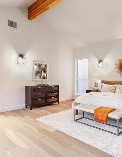 A spacious bedroom with a large bed, wooden furniture, and a decorative mirror. Neutral colors and natural light enhance the modern design.