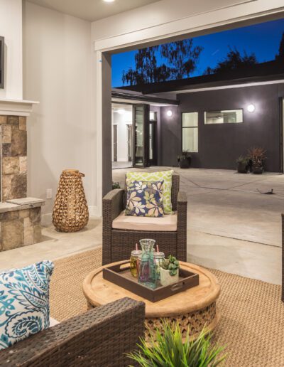 Modern patio with wicker furniture and cushions near a fireplace. Background shows an open garage and a lit room with a bed, under a clear evening sky.