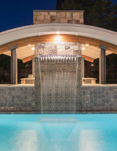 A poolside covered structure with a stone waterfall feature, illuminated at night, surrounded by trees and a stone patio.