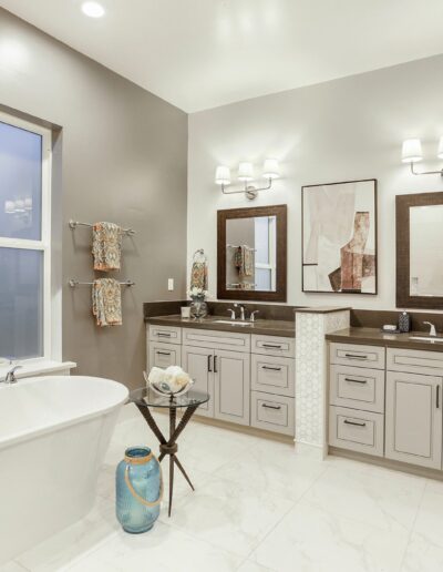 Spacious bathroom with gray double vanity, large mirrors, and modern light fixtures. Freestanding bathtub next to a small side table with decorative vases. Large window and open shelving in view.