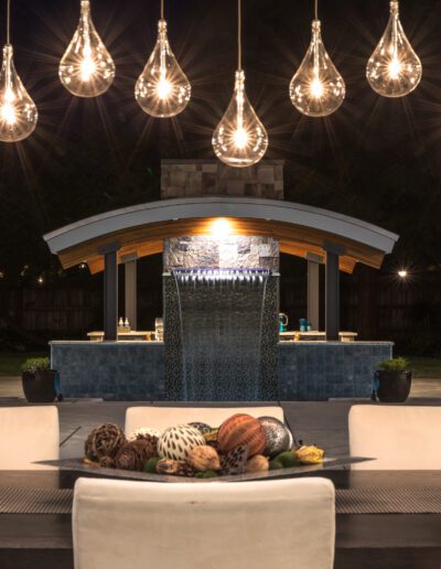 Outdoor dining area with hanging teardrop lights, a table setting with decorative items, and a water feature in the background.