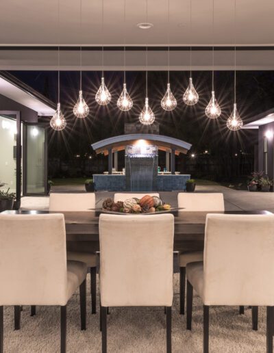 A modern dining room features a dark wooden table with white chairs, pendant lights hanging above, and glass doors opening to an outdoor kitchen area.