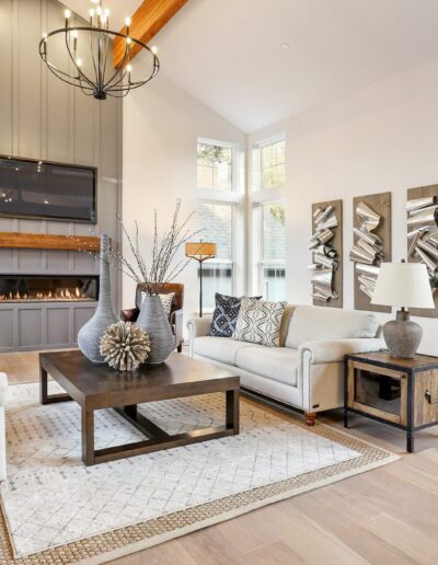 Spacious living room with high ceiling, modern fireplace, neutral-toned furniture, and decorative elements. Large windows allow natural light. Contemporary artwork on the wall.