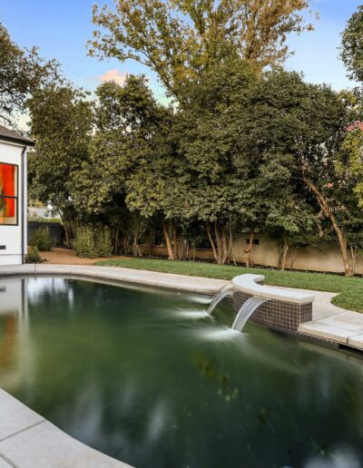 Backyard with a small pool and waterfall feature, next to a modern house with large windows. Surrounded by trees and a lawn.