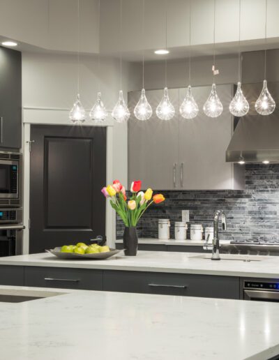 Modern kitchen with gray cabinets, stainless steel appliances, and a white marble island. A vase of tulips and a bowl of green apples decorate the countertop. Pendant lights hang above.