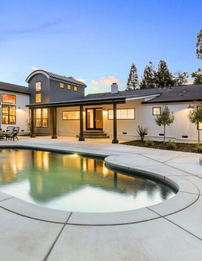 Modern house with large windows, a curved swimming pool in the foreground, and a spacious patio. Surrounded by trees, the house is lit warmly against a twilight sky.