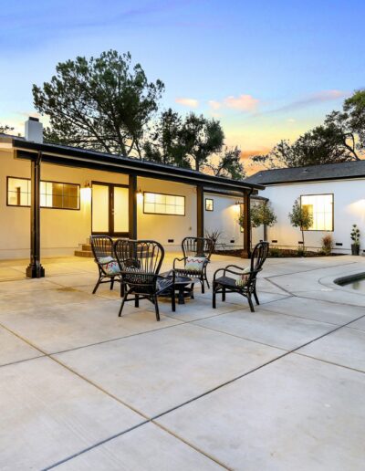 Modern house exterior with large patio, outdoor seating, and a pool. Trees in the background at sunset.