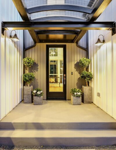 Modern house entrance with a glass door, overhang with lights, and potted plants on either side. Steps lead up to the door.