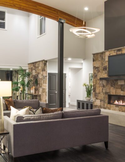 Modern living room with a stone fireplace, wall-mounted TV, gray sofas, and large pendant lights. Wooden ceiling beams and large windows enhance the spaciousness.