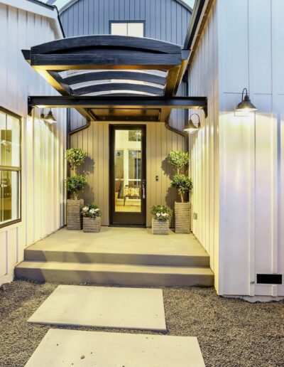 Modern house entrance with a glass door, metal awning, and potted plants on either side. Wood and metal siding with a wooden fence complete the exterior.