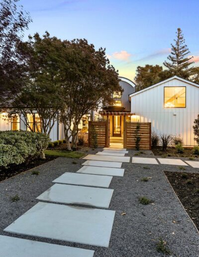 A modern house with a well-lit entrance, surrounded by trees and landscaping, and a stone pathway leading to the front door at dusk.