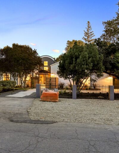 A suburban home with a modern design, surrounded by trees and a fenced front yard, is seen at dusk with soft lighting. A driveway and street are visible in the foreground.