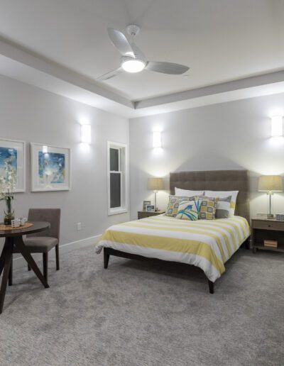 A modern bedroom with a bed, two chairs, a round table, and wall art. The room features a ceiling fan and soft lighting.