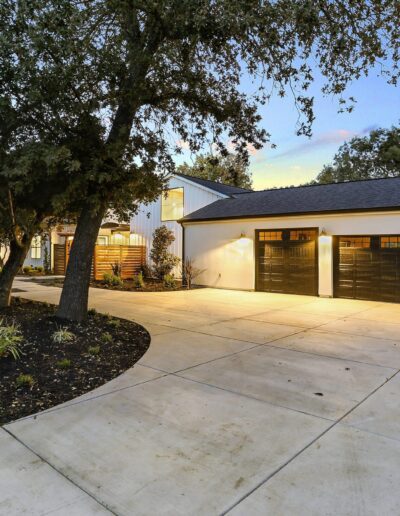 Modern house with a white exterior and a three-car garage, surrounded by trees and landscaped yard, under a twilight sky.