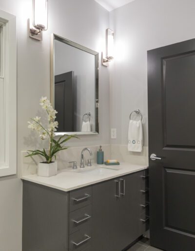 A modern bathroom with a dark vanity, large mirror, and white countertop. There are two wall lights above the mirror and a potted orchid on the counter.