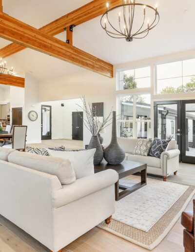 Spacious living room with high ceilings, large windows, and wooden beams. Features white sofas, dark chairs, a central coffee table, and chandeliers. Kitchen area visible in the background.