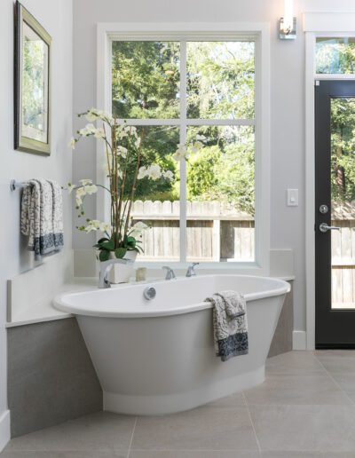 A bathroom features a freestanding bathtub with towels draped over it, a large window with a garden view, and a decorative plant beside the tub.