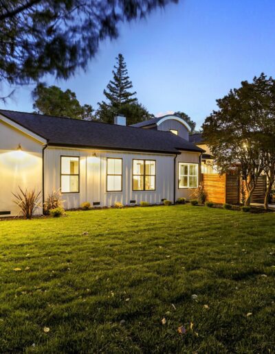 A modern one-story house with large windows, surrounded by a landscaped lawn and trees, is illuminated in the evening.