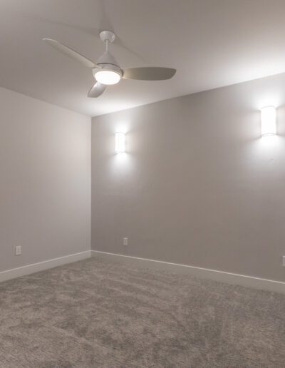 An empty room with gray carpet, two wall lights, a ceiling fan, a window, and a closed door.