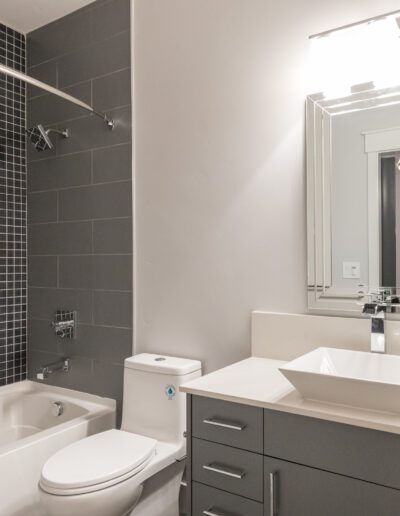 Modern bathroom with a bathtub, black tile wall, white toilet, large mirror, and sink atop a gray vanity.