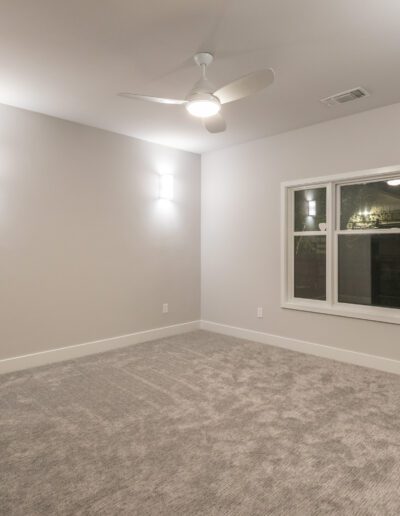 Empty room with gray carpet, white walls, ceiling fan, and two wall lights. A large window overlooks the outside.