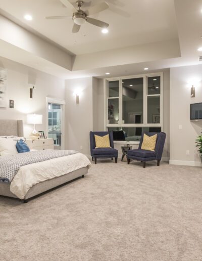 A spacious modern bedroom with a large bed, two chairs, ceiling fan, artwork, and sliding glass doors leading to a patio.