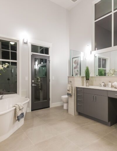 Modern bathroom with a large bathtub, double vanity, chair, and decorative plants. The floor is tiled, and there's a glass shower and high windows.