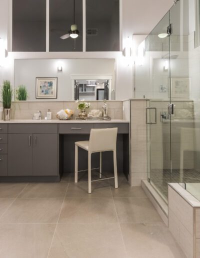 Modern bathroom with large glass shower, gray vanity with chair, dual mirrors, and decorative wall art. Neutral tones create a clean and spacious look.