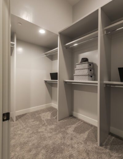 A walk-in closet with gray shelves and clothes rods, beige carpet, and two black boxes and two patterned boxes on the shelves.