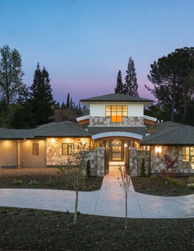 Modern single-story house with large windows, surrounded by trees and a winding driveway, at dusk.