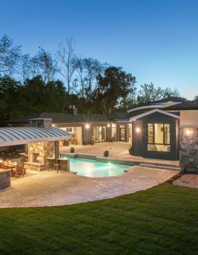 Modern house with a pool and outdoor kitchen under a covered patio, surrounded by trees and lawn, at dusk.