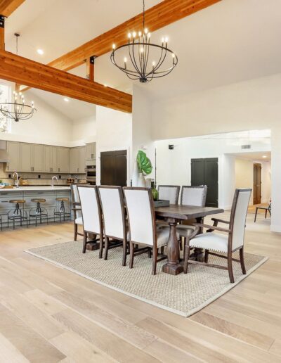 Spacious open-plan kitchen and dining area with light wood flooring, wooden beams, chandeliers, a central island with stools, and a large dining table with white chairs.