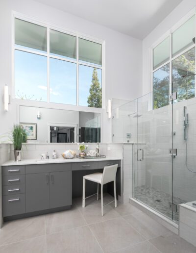 A modern bathroom with a large glass-enclosed shower, a gray vanity with a mirror, and a white chair. Two windows above provide natural light.