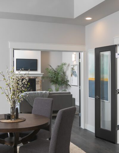 Modern dining and living area with dark furniture, a large window, and a wine storage room. Neutral tones, hardwood floors, and a view into the living room with a couch and fireplace.