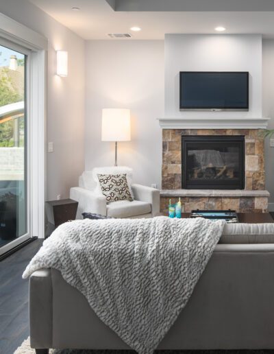 Modern living room with a gray sofa, a stone fireplace, a TV, and a large plant. Glass doors open to a patio outside. A cozy chair and a floor lamp are near the fireplace.