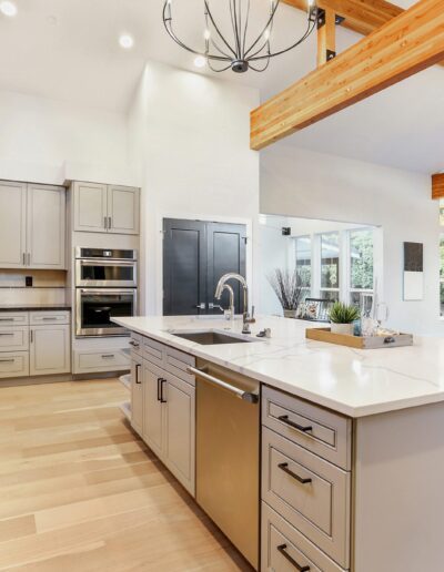 Spacious modern kitchen with light wood flooring, gray cabinets, a large white island, and exposed wooden beams. Dining area visible in the background with large windows.