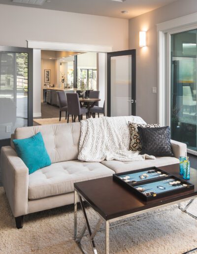 Modern living room with a beige sofa, teal and black cushions, a cozy throw, and a backgammon board on a table. French doors open to a spacious dining area and patio.