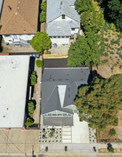 Aerial view of a residential neighborhood showing houses, driveways, lawns, and a dry plot of land with trees and shrubs.