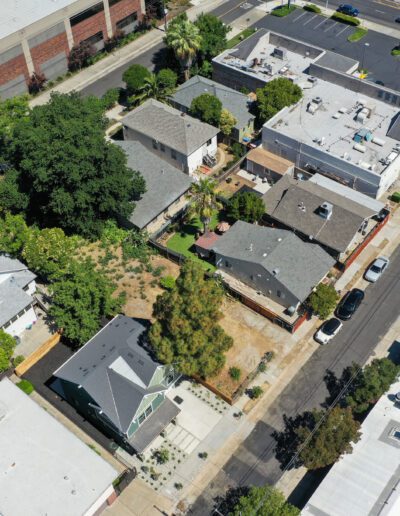 Aerial view of a residential area with houses, trees, and streets. A larger industrial building is visible at the top left. Cars are parked along the streets.