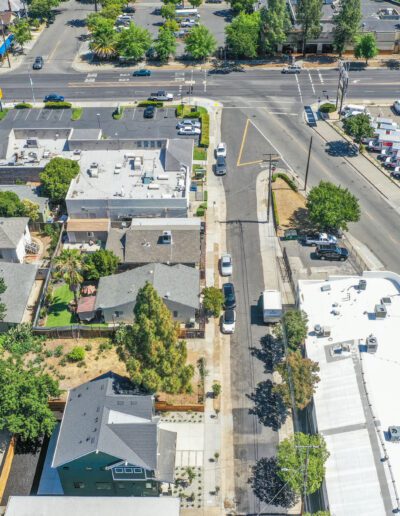 Aerial view of a suburban intersection with residential houses, trees, and parked cars. Commercial buildings and a parking lot are visible in the background.