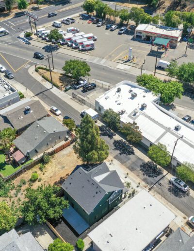 Aerial view of a suburban neighborhood with houses, streets, parked cars, and a small shopping area. Trees are scattered throughout the scene.