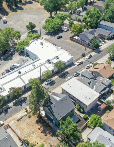 Aerial view of a residential neighborhood with various houses, trees, parked cars, and a white-roofed building at the center.