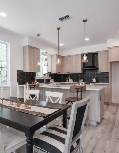 Modern kitchen and dining area with light wood cabinets, black backsplash, a central island, hanging lights, and a dining table with striped chairs. Bright window and sliding door visible.