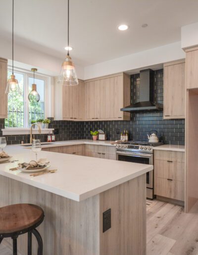 Modern kitchen with light wood cabinets, black tile backsplash, and an island with bar seating. Pendant lights hang above the island. There's a stove with a hood and a sink by the window.