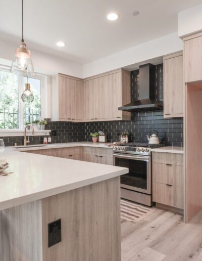 Modern kitchen with light wood cabinets, black backsplash, and white countertops. Features an island with barstools, hanging lights, and a stainless steel stove with range hood.