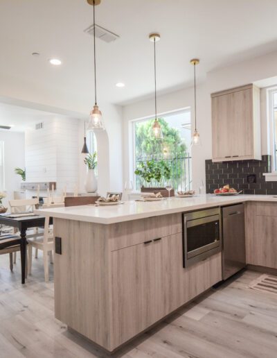 Modern open-concept kitchen and living area with light wood cabinets, pendant lights, and large windows. Dining table set for four and gray couch in the living room.