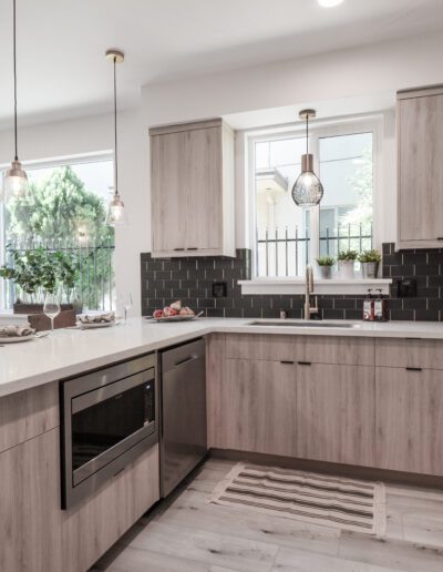 Modern kitchen with light wood cabinets, dark backsplash, and white countertops. Features a built-in microwave, stove, and pendant lights. Large window and dining area visible.